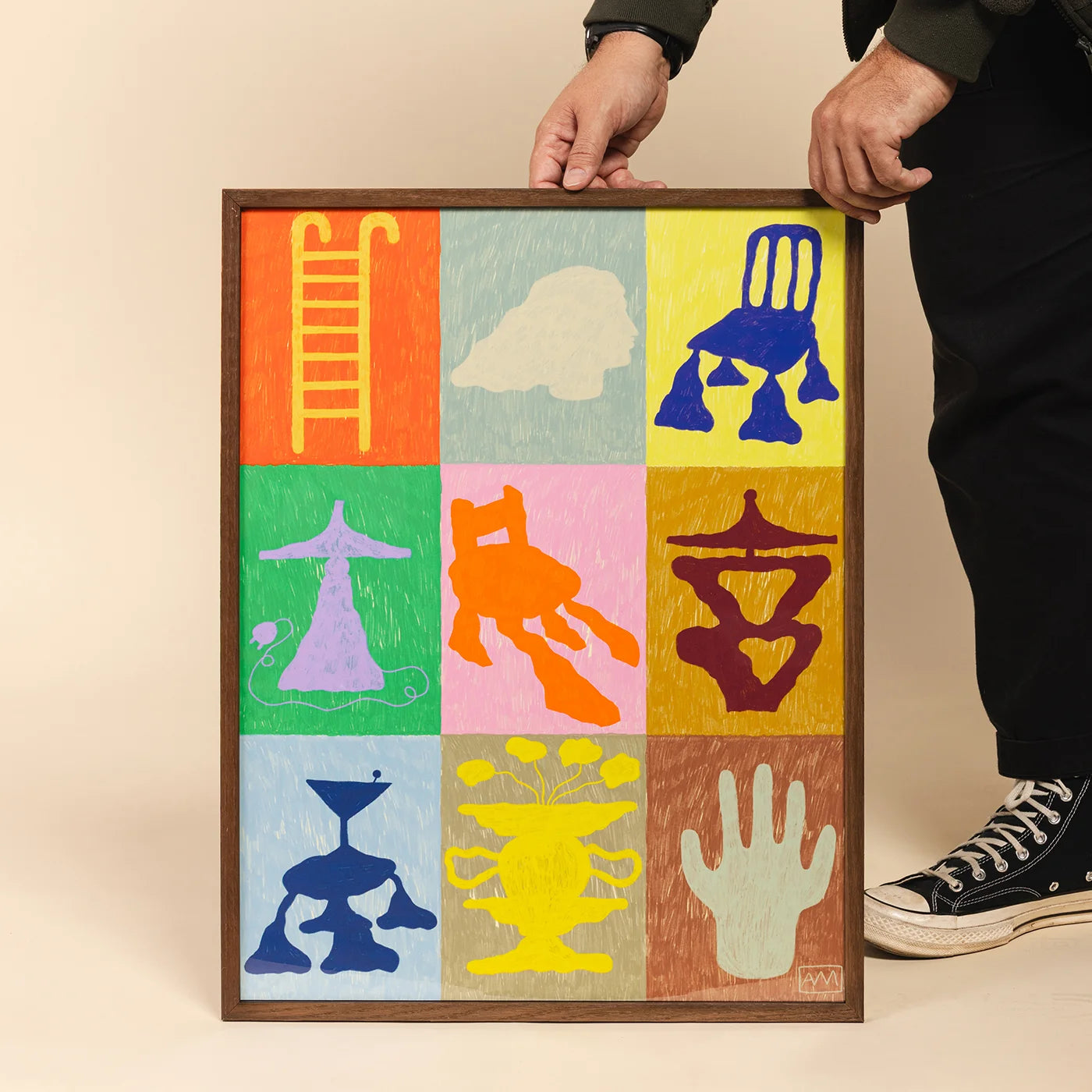 A framed artwork held by a person, showing a 3x3 grid of colorful abstract symbols and shapes on a beige background. 