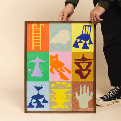 A framed artwork held by a person, showing a 3x3 grid of colorful abstract symbols and shapes on a beige background. 