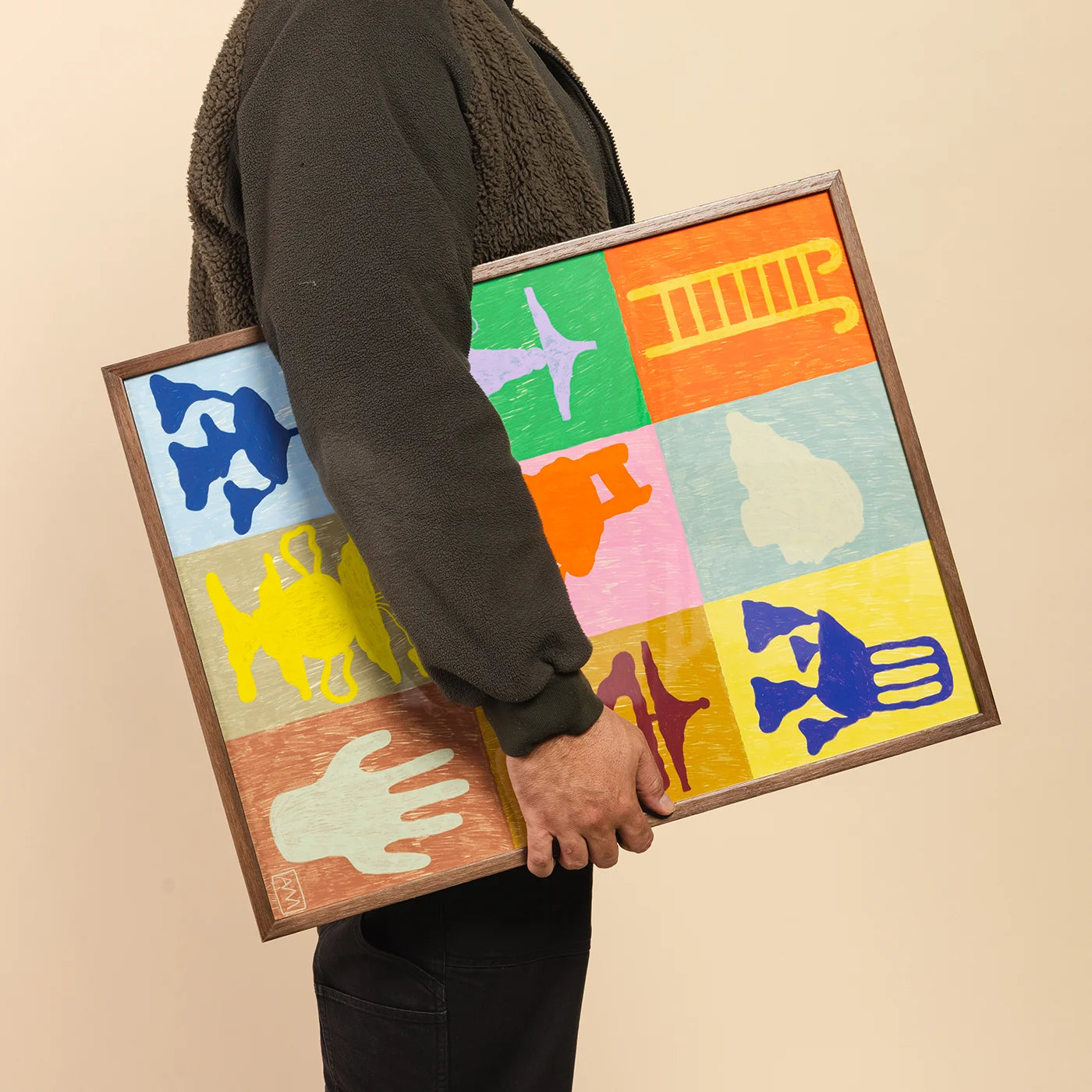 A person in a dark brown fleece jacket holds a square framed art print featuring a colorful 3x3 grid of abstract hand and body shapes in blue, green, orange, pink, yellow, and beige on a warm beige background.