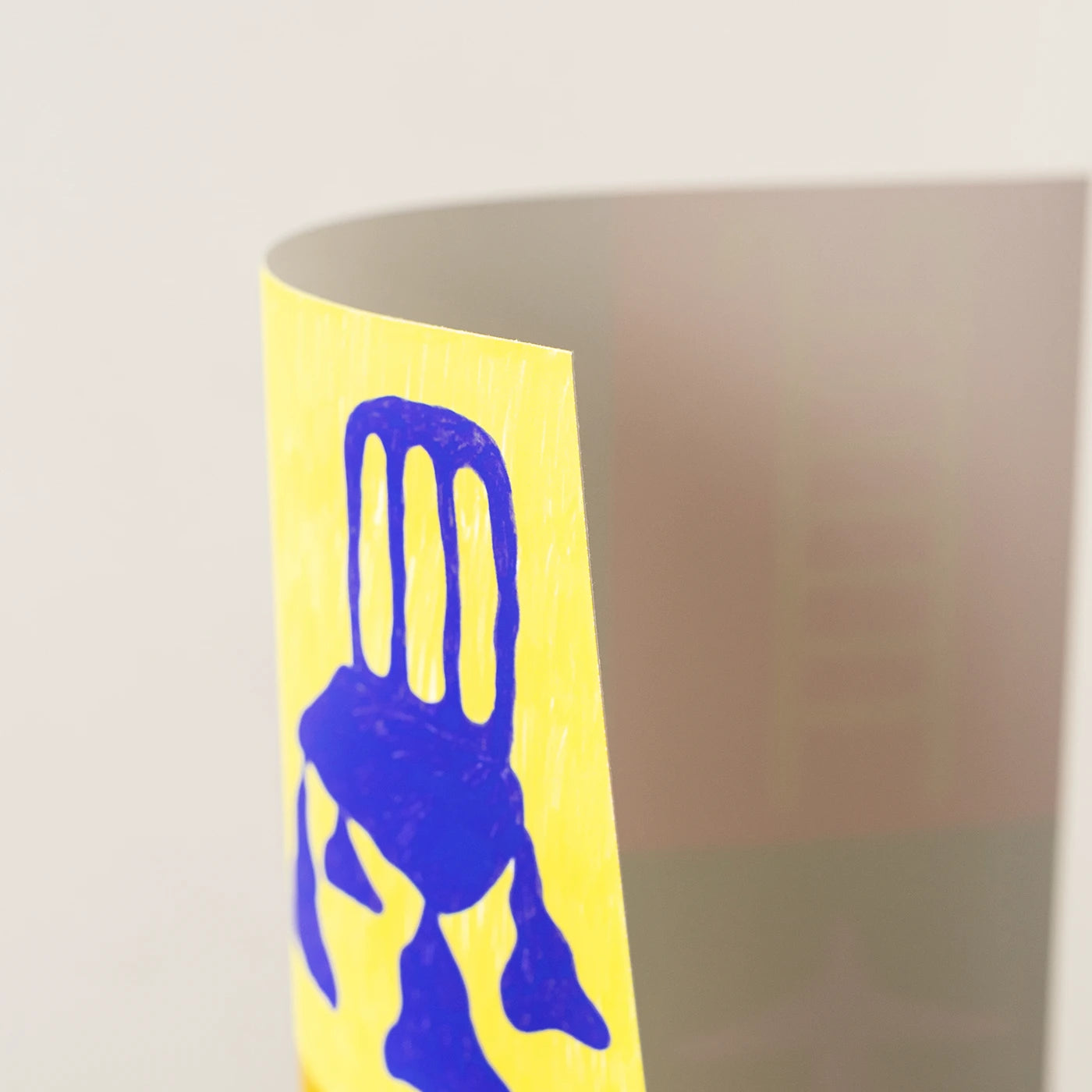 A close-up of yellow wall art with a bold blue chair-like graphic printed on it, photographed against a soft light gray background.