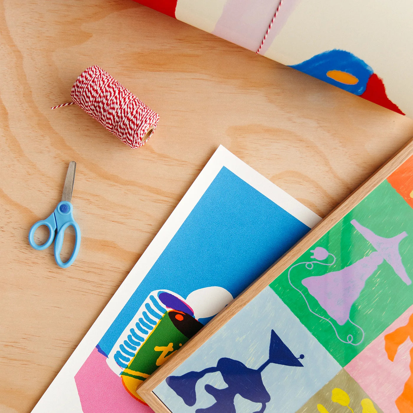 A styled flat-lay of colorful art prints, a roll of red and white bakers twine, blue-handled scissors, and a wooden board on a light wood tabletop.