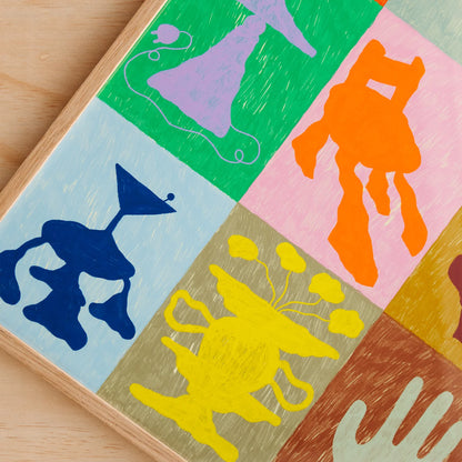 A close-up of a colorful wall art print showing abstract hand-drawn silhouettes in green, pink, blue, yellow, brown, and cream, arranged in a grid on a light wood surface. Framed in a light oak timber frame.