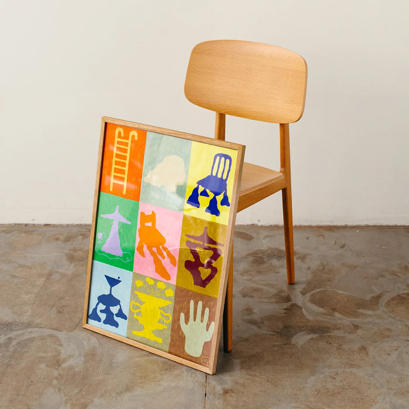 A framed colorful art print leaning against a wooden chair in a minimalist room with a concrete floor and off-white wall.