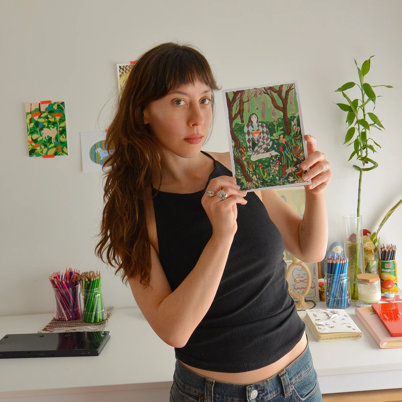 Argentinian artist Yamaha Yjilioff holds up a square art print featuring a colorful forest illustration. She is wearing a black tank top and blue jeans, standing in a bright room with a white desk, art supplies, and a potted plant in the background.