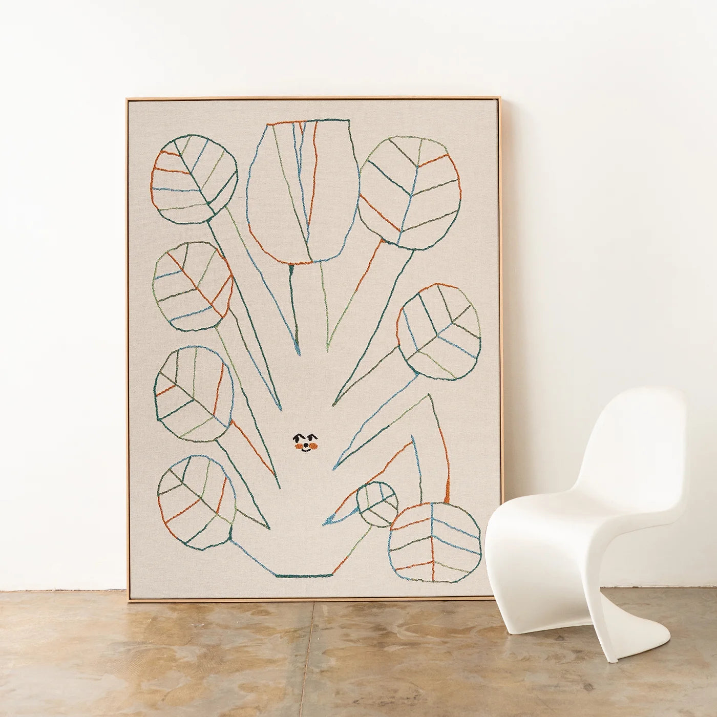 Stick figure inspired abstract artist-designed woven throw with a twig and flower design in colourful linework, framed with an oak timber frame on a white wall with a modern curvy white chair. Woven throw by Slowdown Studio.