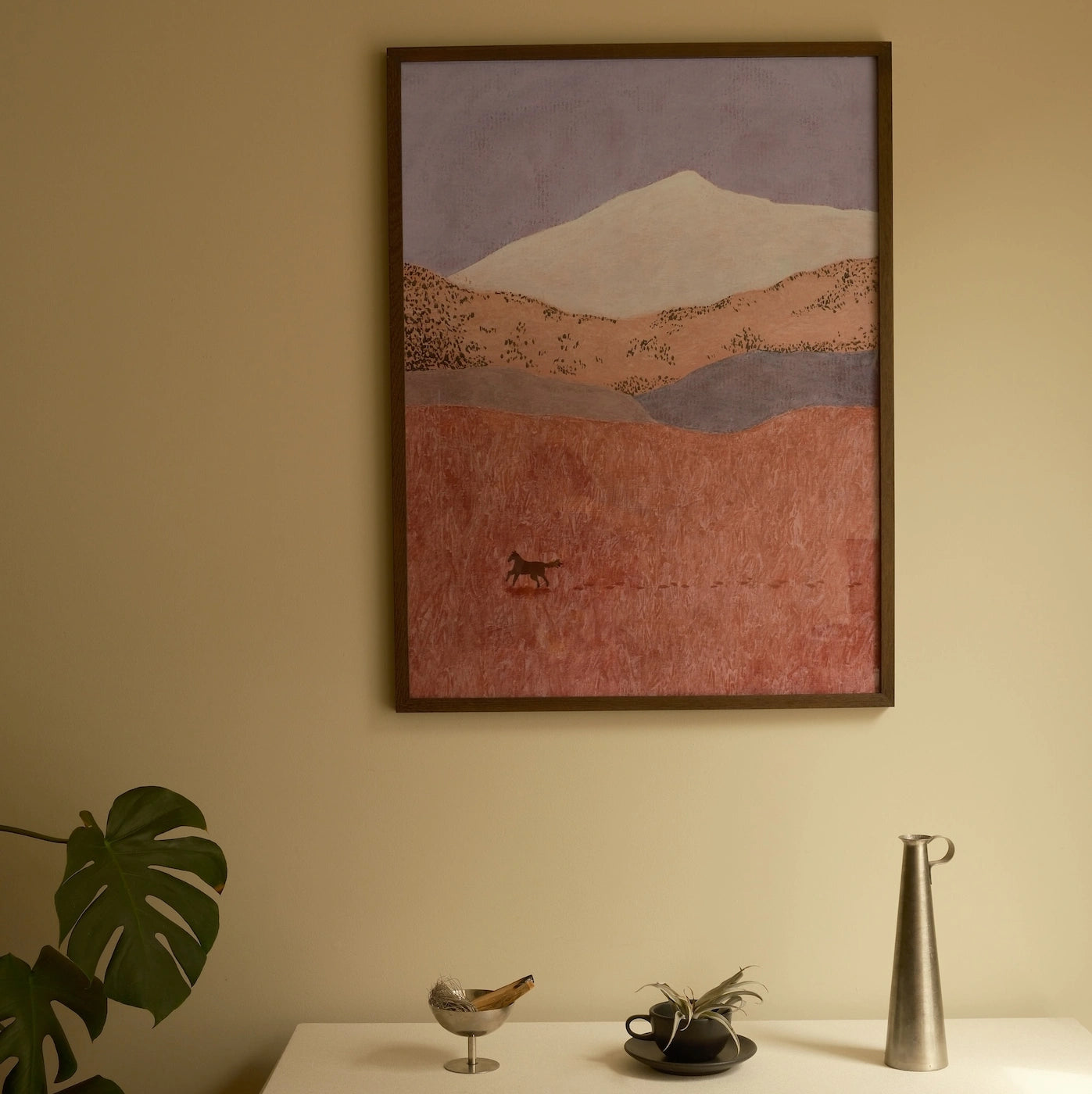 A framed landscape art print featuring a mountain and textured fields in muted pink, brown, and lavender tones, displayed on a beige wall above a white table with decorative objects.