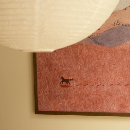 A framed art print featuring a stylized landscape with a small dark horse silhouette on a textured pink field, shown hanging on a beige wall under a white paper lantern.