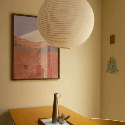 A warm beige dining room corner featuring a large white paper lantern pendant light hanging above a wooden table. On the wall is a framed abstract landscape print with pink, purple, and beige tones, alongside a small turquoise wall decor piece.