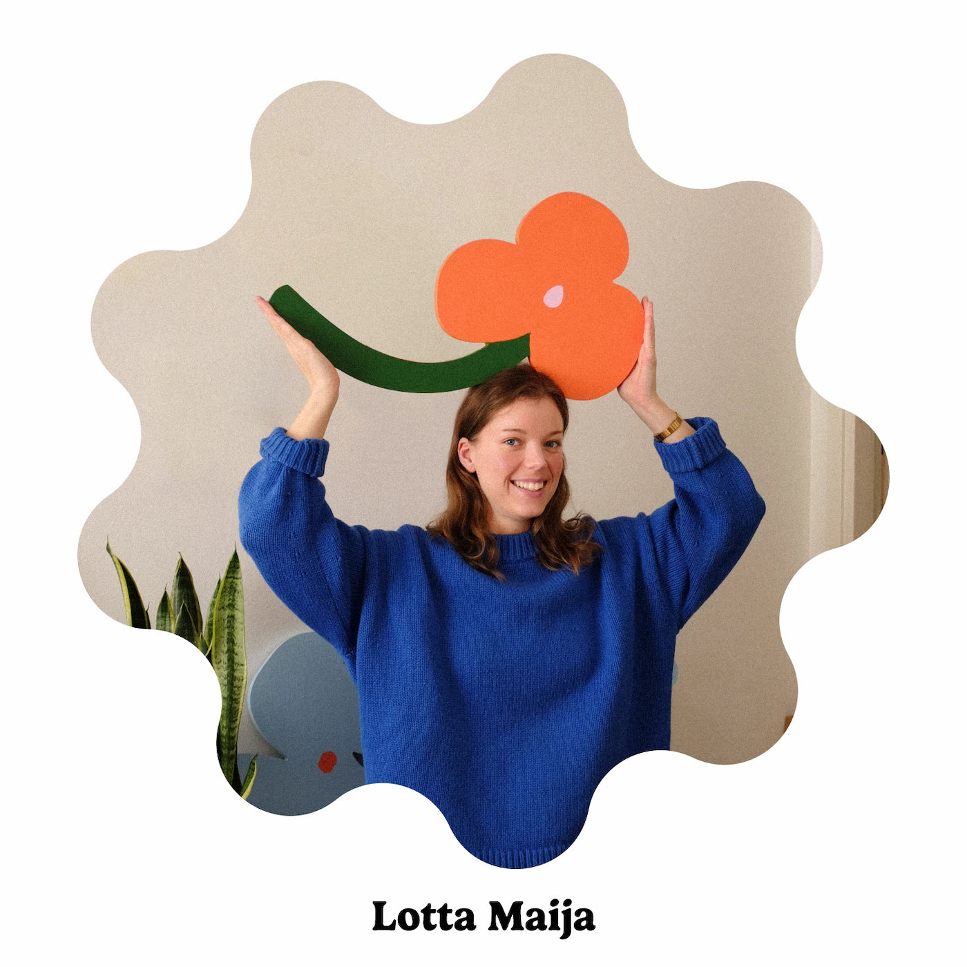 Portrait of Finland-based artist Lotta Maija wearing a bright blue knit sweater holds up a large orange flower decoration with a green stem. The photo is framed inside a wavy scalloped beige shape, with the text &